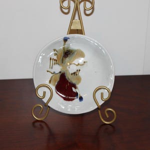 May include: A small white ceramic dish with a brown, red and blue abstract design. The dish is on a gold metal stand.