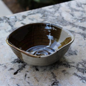 May include: A handmade ceramic bowl with a square shape and a brown and cream color scheme. The interior is a dark brown, with a cream-colored exterior and a brown rim. The bowl has a glossy finish.