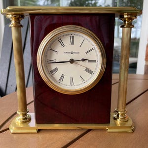 May include: A Howard Miller clock with a cream-colored face, Roman numerals, and gold-tone hands. The clock is encased in a rectangular, dark wood frame with gold accents and decorative columns. The clock's face reads "HOWARD MILLER".