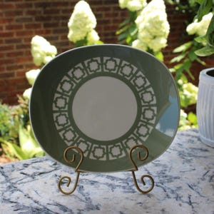 May include: A green and white decorative plate with a geometric pattern, displayed on a gold-colored metal stand. The plate has a central white circle and a border of repeating shapes. The background includes greenery and a brick wall.