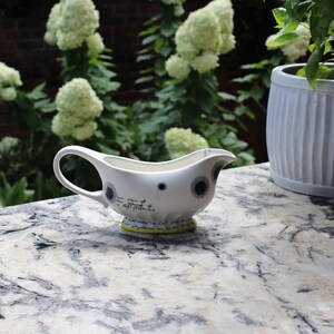 May include: A white ceramic gravy boat with a handle and a spout. The boat features a yellow base and is decorated with blue and black circular patterns and script. The gravy boat is on a marble surface, with greenery in the background.
