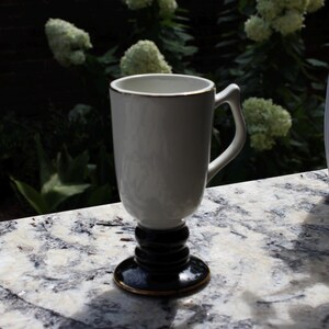 May include: A white ceramic mug with a gold rim and handle. The mug sits on a black pedestal base with a gold trim. The mug is on a marble surface with green foliage in the background.