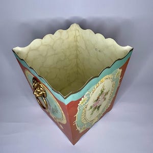 May include: A square, red, white, and blue planter with a scalloped rim and a floral design. The planter has a gold lion head handle.