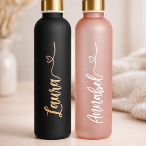 May include: Two personalized water bottles. One is black with gold lettering that says "Laura". The other is pink with white lettering that says "Annabel". Both bottles have gold-colored lids.