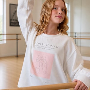 May include: A white long-sleeve sweatshirt featuring a graphic print of ballet slippers on a light pink square. The text reads LE COURS DE DANSE, MUSÉE D'ORSAY À PARIS, une collection impressionniste, DEGAS, and 1834-1917 in black lettering.