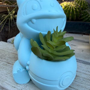 Könnte beinhalten: Hellblauer Bulbasaur-Pflanztopf mit einer grünen Sukkulente. Der Pflanztopf ist ein detailliertes 3D-Druckdesign des beliebten Pokémon-Charakters. Der Pflanztopf steht auf einer Holzoberfläche.