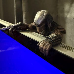 Puede incluir: Figura detallada de un personaje Predator, con un casco marrón y plateado texturizado y rastas. La figura está posada con las manos apoyadas en una superficie, sosteniendo un arma. La figura es una representación detallada del personaje de ciencia ficción.