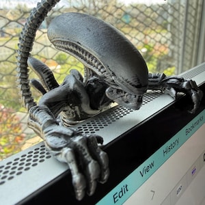 Pode incluir: Figura cinzenta detalhada de um Xenomorfo com uma longa cauda segmentada e mãos com garras. A criatura está pousada na borda de um monitor de computador, com a cabeça e a cauda arqueadas para cima. O monitor exibe as palavras "File", "Edit", "View", "History" e "Bookmarks".