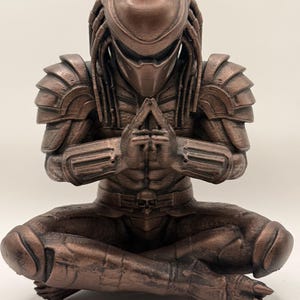 May include: A bronze-colored Predator figurine in a seated yoga pose. The figure has detailed armor, dreadlocks, and a mask. Hands are clasped in a prayer position. The figurine is a unique blend of sci-fi and spiritual themes.
