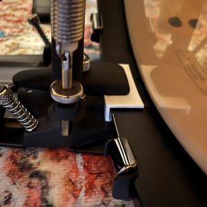 以下が含まれることがあります： 銀色のバネと金具が付いた黒いドラムペダルのクローズアップ。ペダルは白いドラムに取り付けられています。画像は、バネや調整可能なコンポーネントなど、ペダルのメカニズムの詳細を示しています。