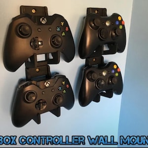 May include: Four black Xbox controller wall mounts are attached to a light blue wall. Each mount holds a black Xbox controller. The text "XBOX CONTROLLER WALL MOUNT" is at the bottom of the image.