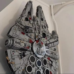 May include: A large, gray and red Lego model of the Millennium Falcon spaceship. The model is built in a detailed and realistic style, with many small parts and intricate details. The model is displayed on a wall, and it appears to be a very large and impressive piece of Lego art.