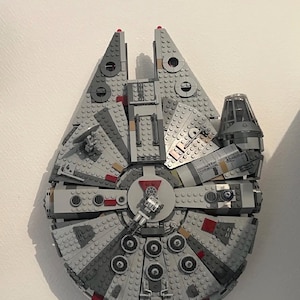 Puede incluir: Modelo Lego gris de la nave espacial Halcón Milenario. El modelo presenta una construcción detallada con varios tonos grises, detalles en rojo y una cabina. La nave espacial es un artículo popular de la franquicia Star Wars.