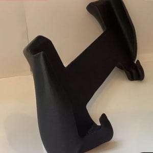 Peut inclure: Support de t&eacute;l&eacute;phone en plastique noir avec deux bras incurv&eacute;s et une base. Le support est con&ccedil;u pour maintenir un t&eacute;l&eacute;phone en position verticale.
