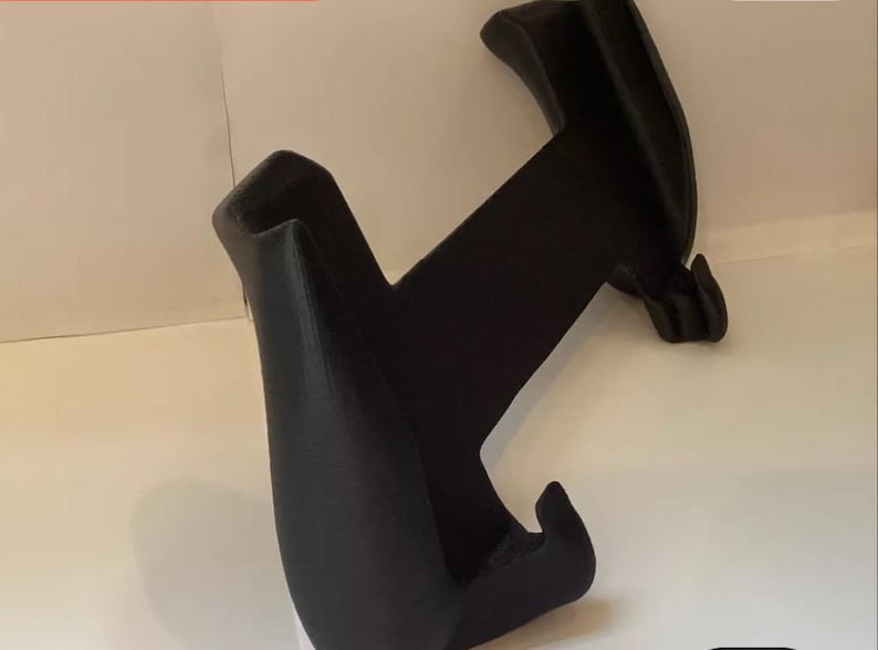 Peut inclure: Support de t&eacute;l&eacute;phone en plastique noir avec deux bras incurv&eacute;s et une base. Le support est con&ccedil;u pour maintenir un t&eacute;l&eacute;phone en position verticale.