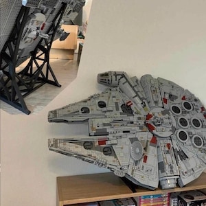 Puede incluir: Un modelo detallado de la nave espacial Halcón Milenario, construido con bloques de construcción entrelazados. La nave gris y blanca presenta detalles intrincados, incluyendo portillas circulares y acentos rojos. Se exhibe en una estantería de madera y un soporte negro.
