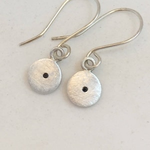 May include: A pair of silver drop earrings. Each earring features a small, brushed silver disc with a single black dot in the center. The earrings have a simple, elegant design with curved ear wires.