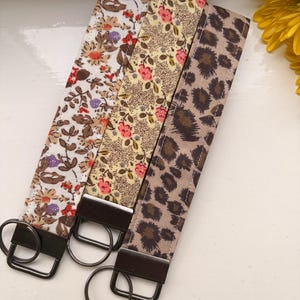 May include: Three fabric wristlet keychains with dark metal hardware. The first features a white background with brown, orange, and purple floral patterns. The second has a yellow background with small pink flowers. The third displays a brown leopard print.