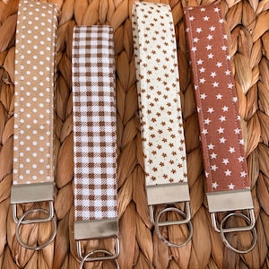 May include: Four fabric wristlet keychains with silver-toned metal hardware and split rings. The first features white polka dots on tan, the second a brown and white gingham check, the third small brown flowers on cream, and the fourth white stars on rust.