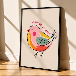 May include: A framed wall art print featuring a stylized illustration of a bird with pink, yellow, orange, and green color blocks. The bird has a black outline and the text "WINGING IT DAILY" is printed in red above the bird on a cream background.