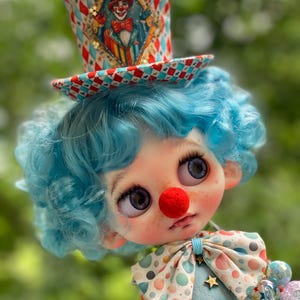 Op de afbeelding: Een pop met krullend lichtblauw haar en een rode neus, met een hoge hoed versierd met een ruitvormige clownillustratie en een rood, wit en blauw harlekijnpatroon. De pop heeft een vlinderdas met stippen en een lichtblauwe outfit met gouden sterren.