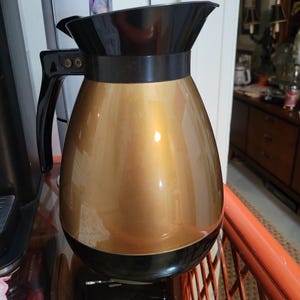 May include: A gold-colored coffee carafe with a black lid and handle. The carafe has a shiny, metallic finish.