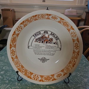 May include: White ceramic Taco Salad serving bowl with an orange floral border and recipe text. The bowl is displayed on a black metal stand. The text includes a recipe for taco salad.