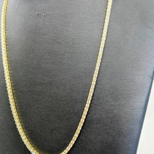 Könnte beinhalten: Eine goldfarbene Tennis-Halskette mit einer durchgehenden Reihe kleiner, klarer Edelsteine. Die Halskette wird auf einer schwarzen Schmuckbüste präsentiert, die ihr Design und ihren Glanz hervorhebt. Die Kette ist ein klassischer, eleganter Stil.