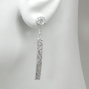May include: Silver dangle earrings with a clear cubic zirconia stone at the top and a long, narrow, silver bar with an intricate design hanging below.