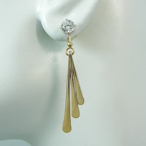 May include: A gold-toned earring with a clear, round gemstone at the top. Three elongated, teardrop-shaped gold pieces dangle below. The earring is displayed against a white background, highlighting its elegant design.