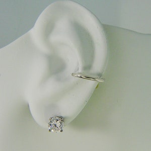 May include: A close-up of a white ear with two pieces of jewelry. A round, clear, sparkling stud earring is in the earlobe. A silver-colored ear cuff is on the upper ear.