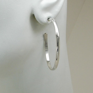 May include: A single, silver-colored hoop earring. The earring is a classic, minimalist design, featuring a smooth, rounded outer edge and a flat, straight post. The earring is shown on a white mannequin head, highlighting its shape and shine.