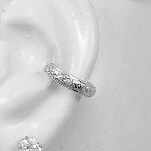 May include: A silver ear cuff featuring an intricate floral pattern. The cuff is positioned on the upper ear, showcasing its detailed, engraved design. The image is a close-up, emphasizing the texture and design of the jewellery.