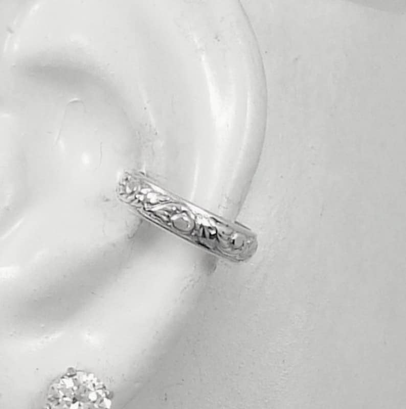 May include: A silver ear cuff featuring an intricate floral pattern. The cuff is positioned on the upper ear, showcasing its detailed, engraved design. The image is a close-up, emphasizing the texture and design of the jewellery.