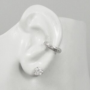 May include: A silver ear cuff featuring a floral pattern and a clear, round stud earring. The ear cuff is positioned on the upper ear, with the stud earring in the earlobe. The jewellery is displayed on a white ear model.
