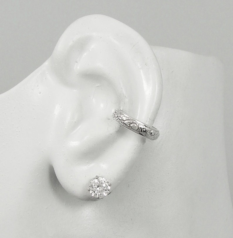 May include: A silver ear cuff featuring a floral pattern and a clear, round stud earring. The ear cuff is positioned on the upper ear, with the stud earring in the earlobe. The jewellery is displayed on a white ear model.