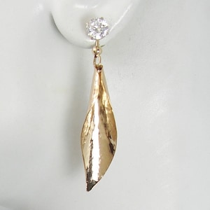 May include: A gold-colored earring with a dangling, leaf-shaped design. The earring features a small, round, clear gemstone at the top. The leaf-shaped element has a textured surface and a polished finish, reflecting light.