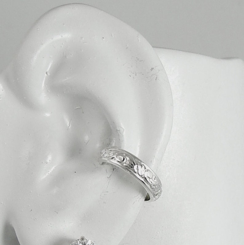 May include: A silver ear cuff with a floral pattern is displayed on a white ear model. The cuff, a band with detailed carvings of flowers and leaves, wraps around the ear cartilage. The jewellery is shown against a plain white background.
