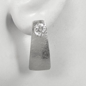 May include: A single silver-toned earring with a brushed metal finish. The earring features a rectangular shape with a rounded bottom and a sparkling, round, clear gemstone set near the top. The earring is shown on a white background.