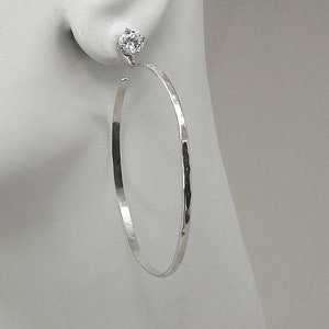May include: A pair of silver hoop earrings with a hammered texture. Each earring features a small, clear, round gemstone stud at the top. The hoops are large and have a simple, elegant design, suitable for everyday wear.