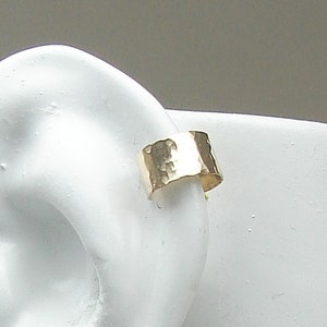 May include: A gold-colored ear cuff with a hammered texture. The cuff is a wide band that wraps around the ear. The jewelry is shown on a white background.
