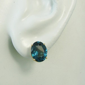 May include: A single gold stud earring featuring an oval-cut, teal-colored gemstone. The earring is set in a four-prong gold setting, showcasing the vibrant color of the stone. The earring is displayed on a white mannequin ear.
