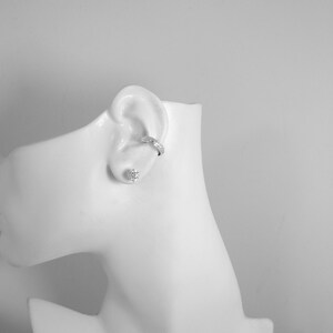 May include: A white mannequin head displaying a silver ear cuff and a stud earring. The ear cuff is worn on the upper ear, and the stud earring is in the earlobe. The jewellery is shown against a white backdrop.