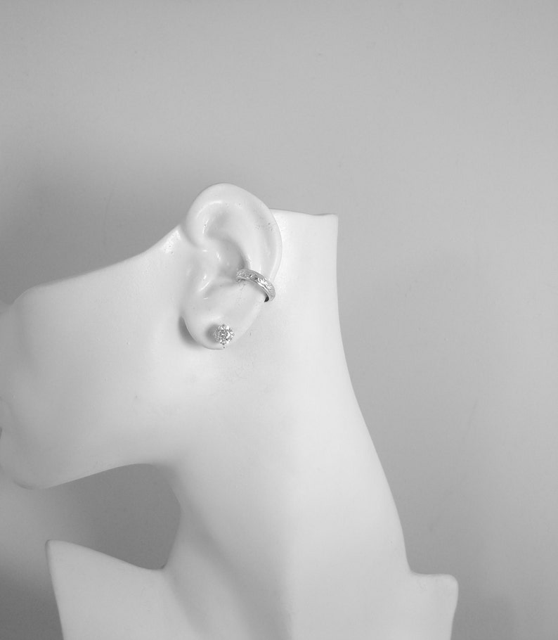 May include: A white mannequin head displaying a silver ear cuff and a stud earring. The ear cuff is worn on the upper ear, and the stud earring is in the earlobe. The jewellery is shown against a white backdrop.