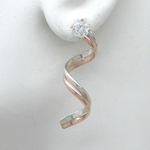 May include: A pair of silver and rose gold earrings with a clear crystal in the center. The earrings are twisted and have a spiral design.