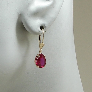May include: A gold-colored earring with a lever back clasp. The earring features a teardrop-shaped, deep red gemstone. The earring is shown on a white mannequin head.