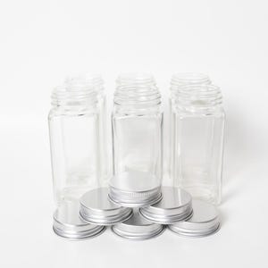 May include: A set of six clear glass spice jars with silver metal screw-on lids. The jars are rectangular with rounded corners. The lids are flat with a ridged edge. The jars and lids are arranged on a white background.