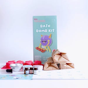 May include: A Kiss Naturals DIY Bath Bomb Kit box featuring a purple monster illustration, three brown paper bags, a red heart-shaped silicone mould, a small metal bowl, three small glass bottles, and a sheet of stickers with the word TAP printed.