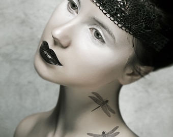 May include: A portrait of a person with pale skin, dark lipstick, and a black lace headband. Two dragonfly tattoos are visible on the neck. The image is in grayscale, with a focus on the face and upper body.