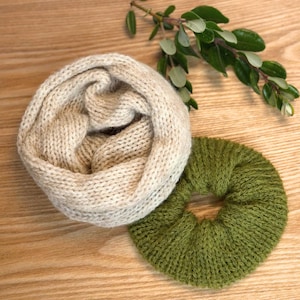 May include: A beige knitted infinity scarf and a green knitted scrunchie rest on a wooden surface. The scarf is a soft, neutral color, and the scrunchie is a vibrant green. A sprig of greenery is in the background.
